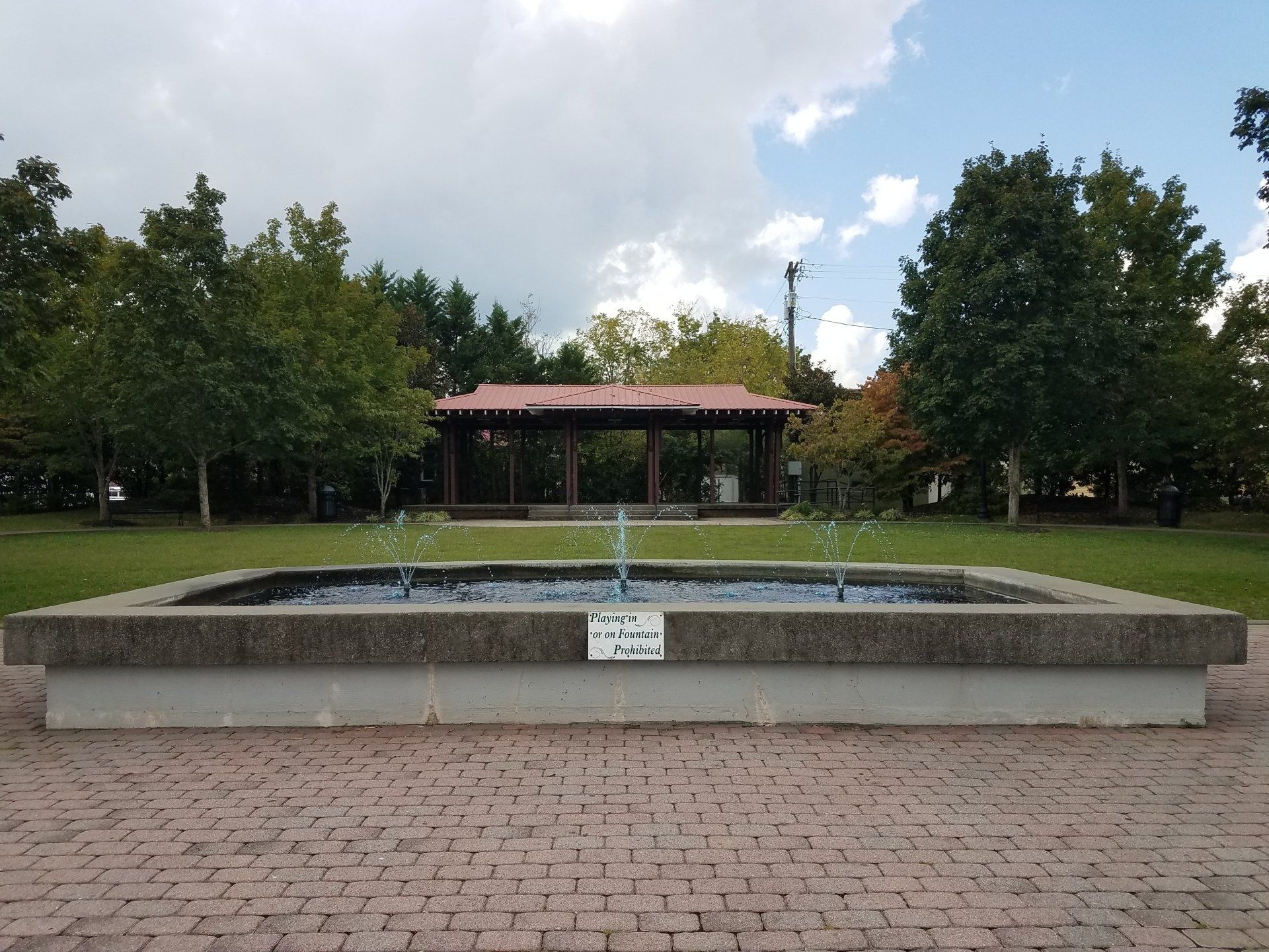 Outdoor fountain in a brick plaza with trees and a pavilion in the background