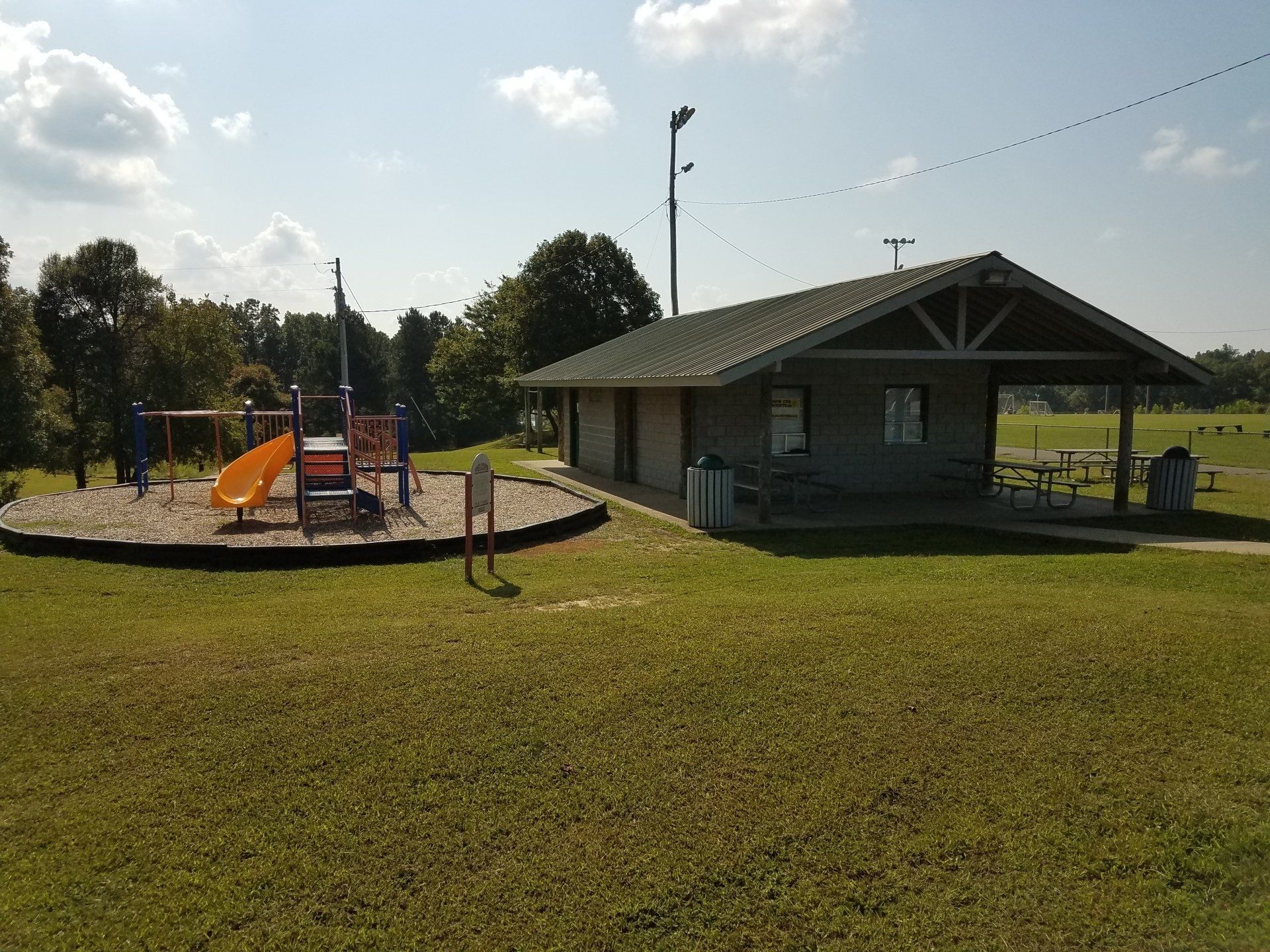 Playground beside a pavilion in a sunny park, with grassy field and trees in the background