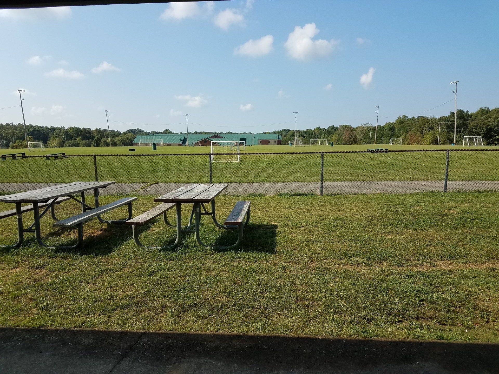 Picnic tables on a grassy field beside a chain-link fence under a clear blue sky.