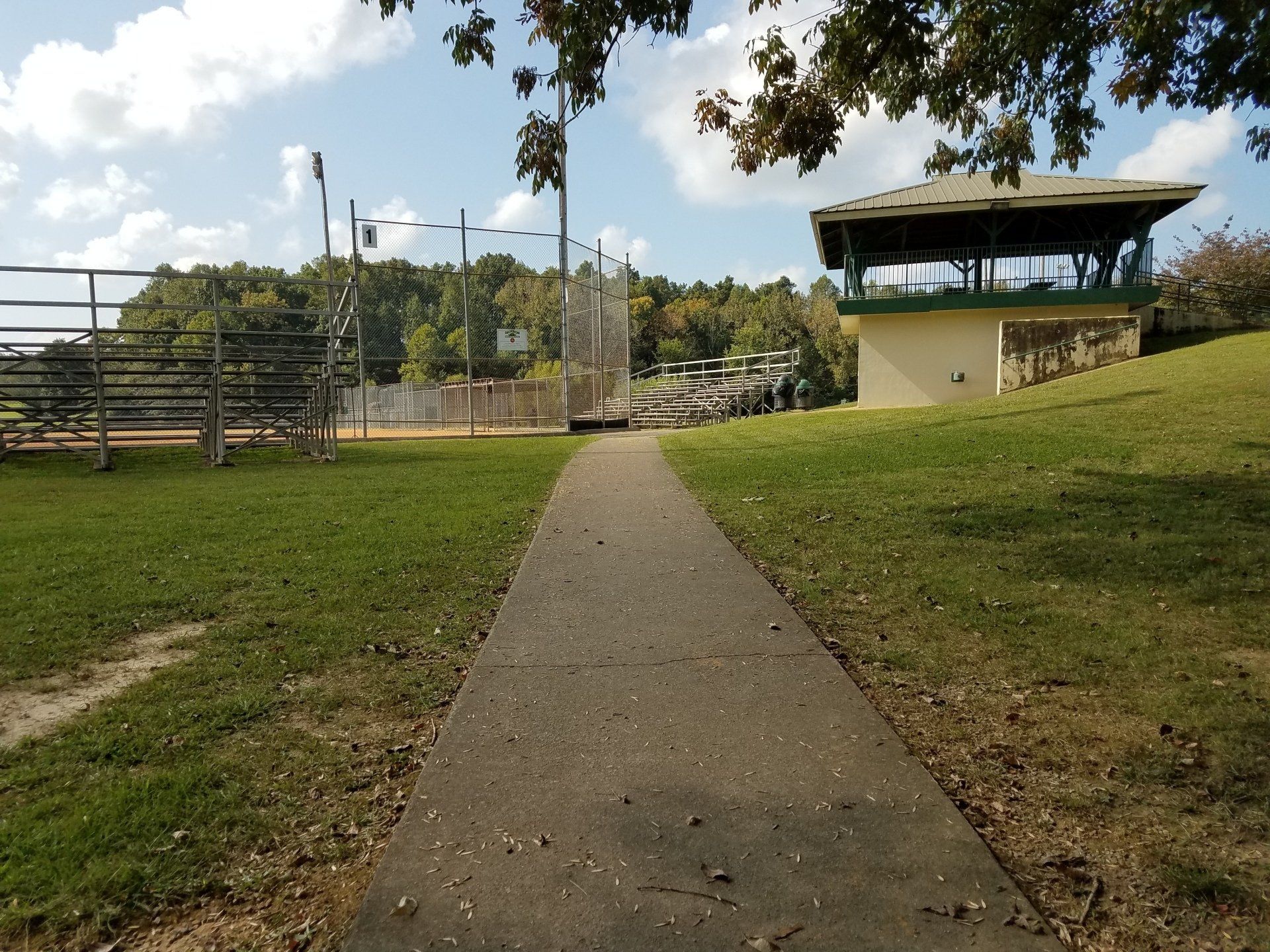Park path leading through grassy area toward a small pavilion and fenced sports field under a cloudy sky