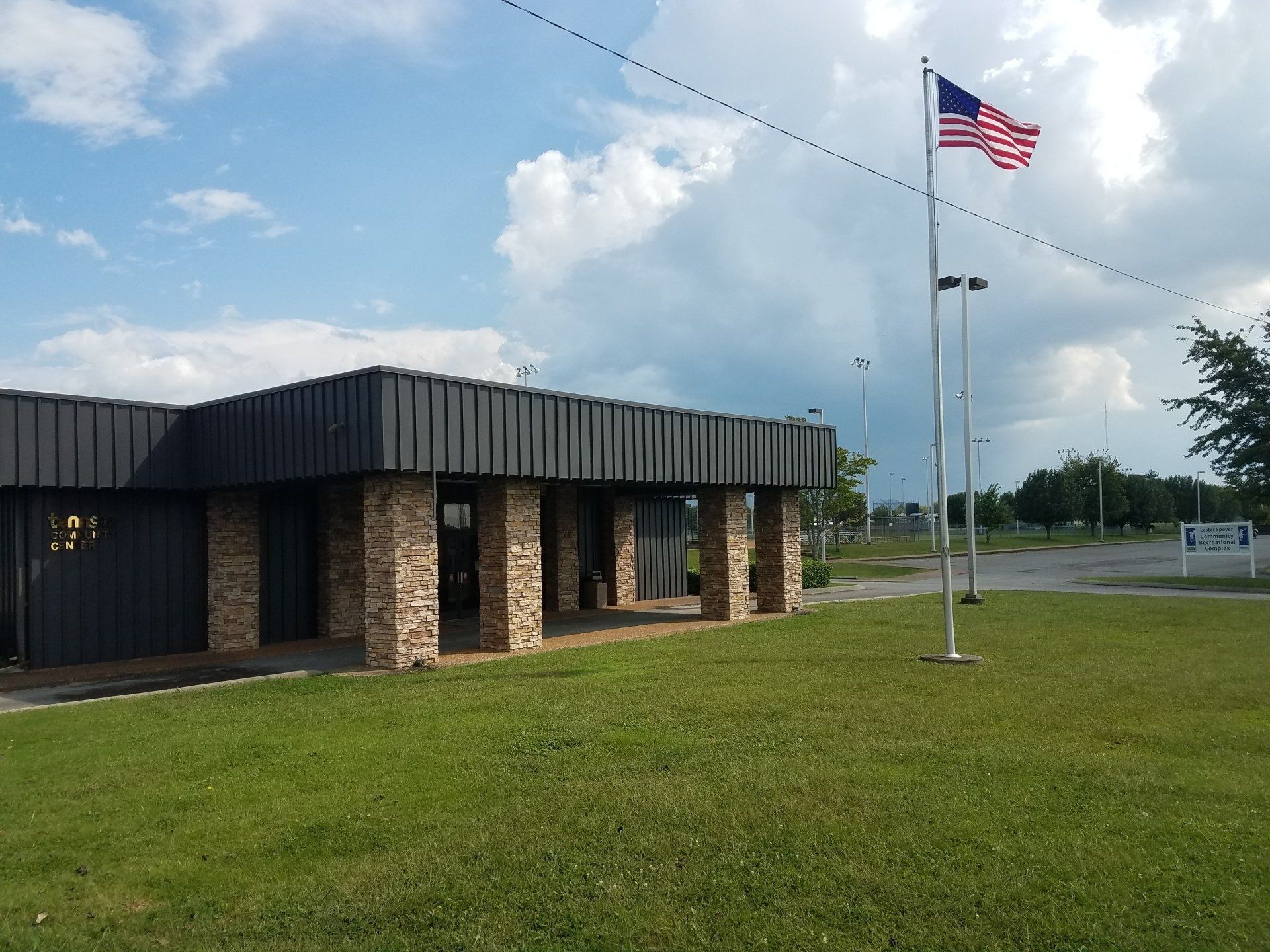 Small brick building with black roof, American flag on a pole, and grassy lawn under a partly cloudy sky