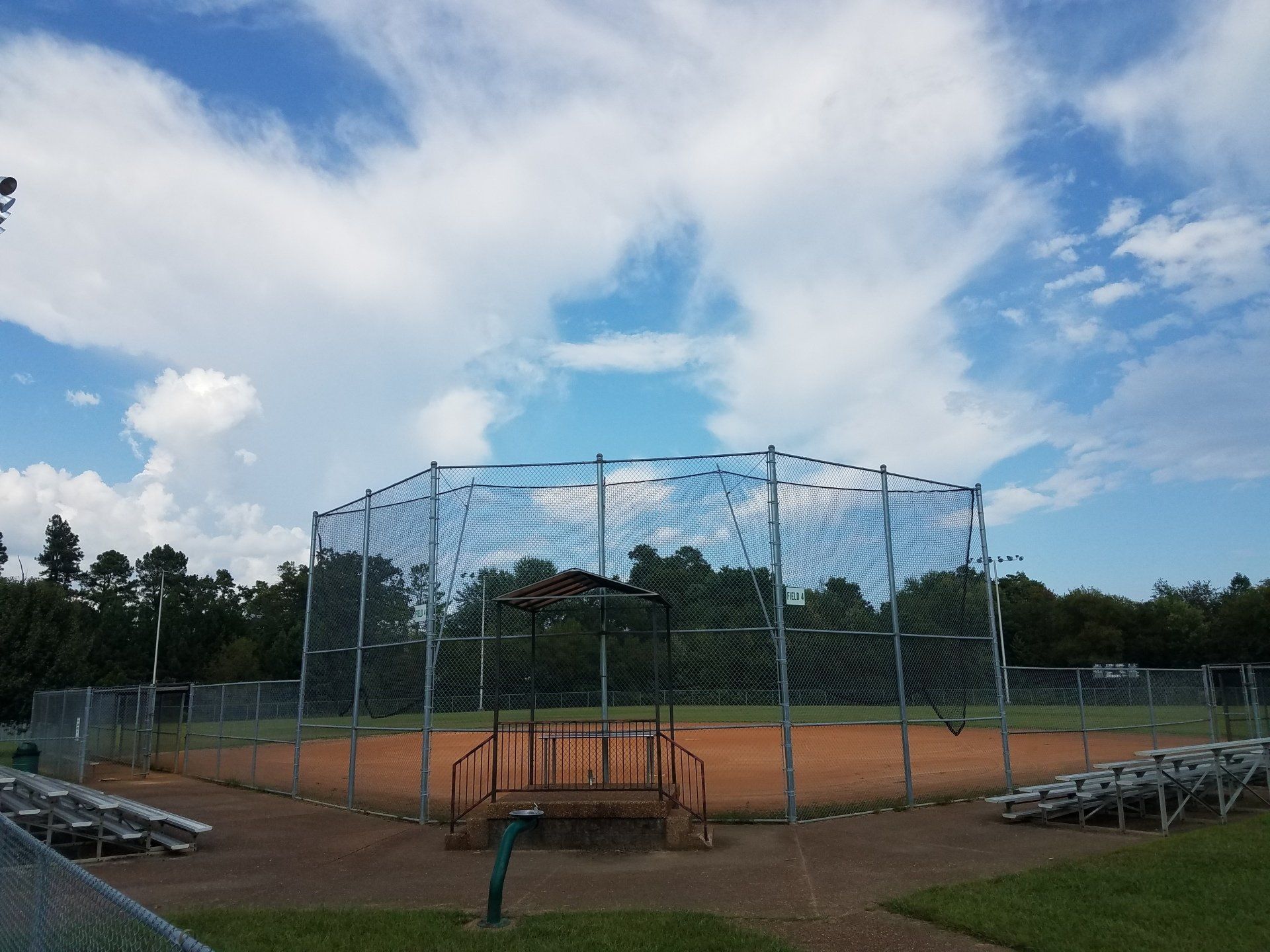 Baseball field with backstop netting, dirt infield, and bleachers under a cloudy blue sky