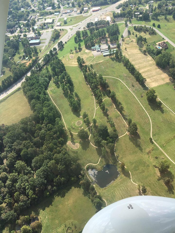 Aerial view of a green golf course with fairways, trees, and nearby roads