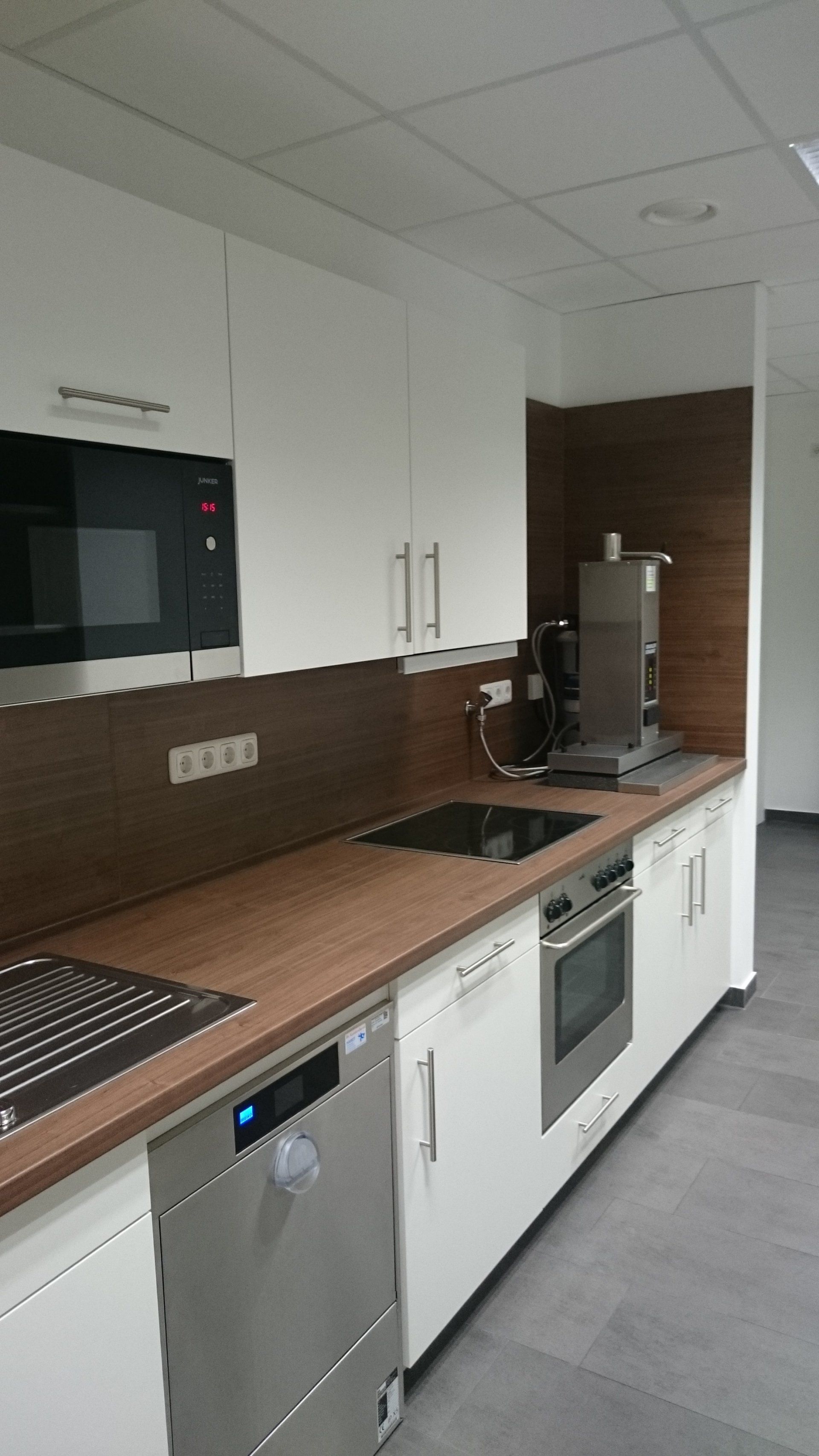Moderne Büroküche mit weißen Schränken, Arbeitsplatten aus Holz, Edelstahlgeräten und einer Kaffeemaschine.
