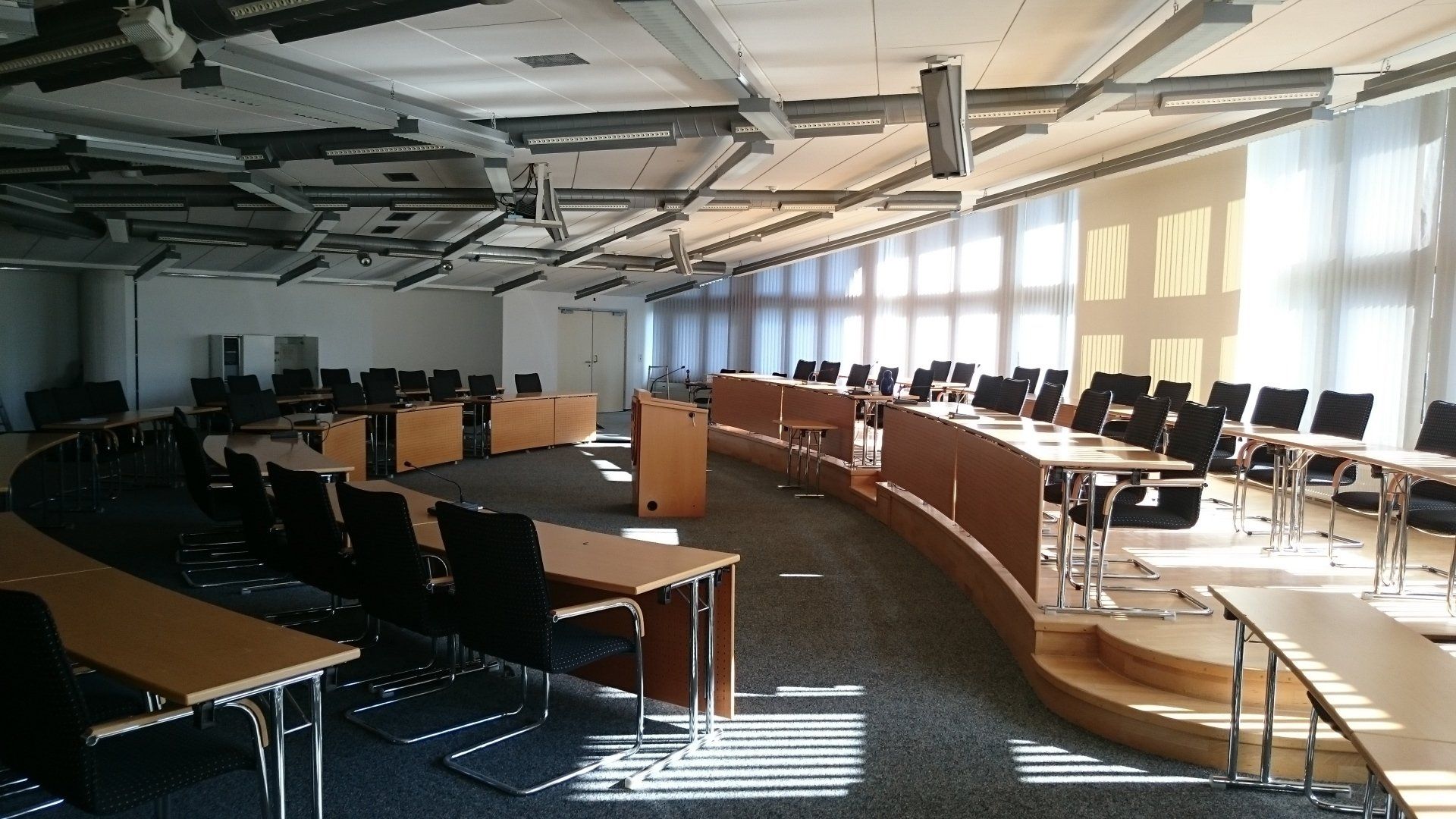 Ein großer, moderner Konferenzraum mit geschwungenen Holztischen, schwarzen Stühlen und hellem Sonnenlicht, 