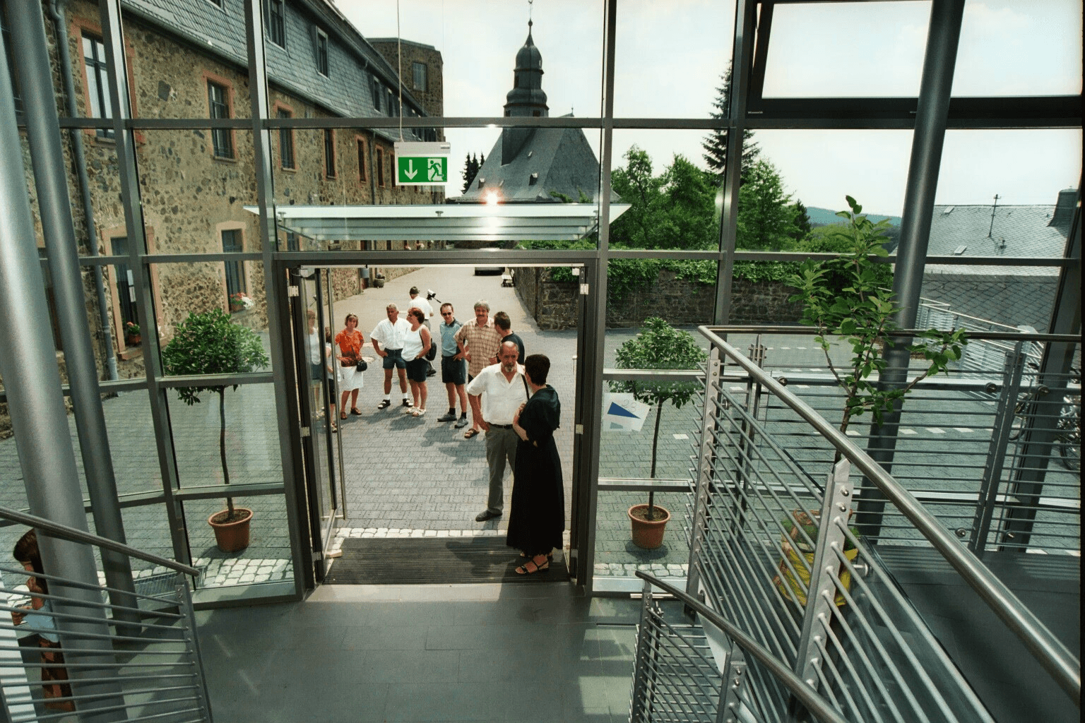 Eine Gruppe von Menschen versammelt sich in einem Steinhof, der von einem Gebäude mit Glaswänden und einer Metalltreppe