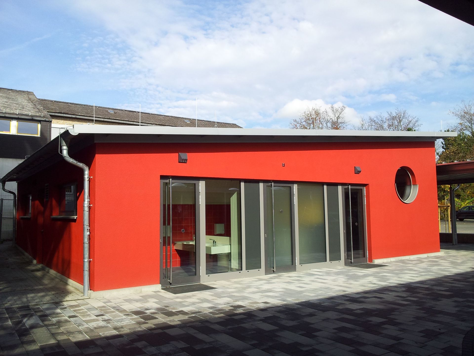 Ein modernes, leuchtend rotes Gebäude mit raumhohen Glastüren und einem runden Fenster auf einem gepflasterten Innenhof.