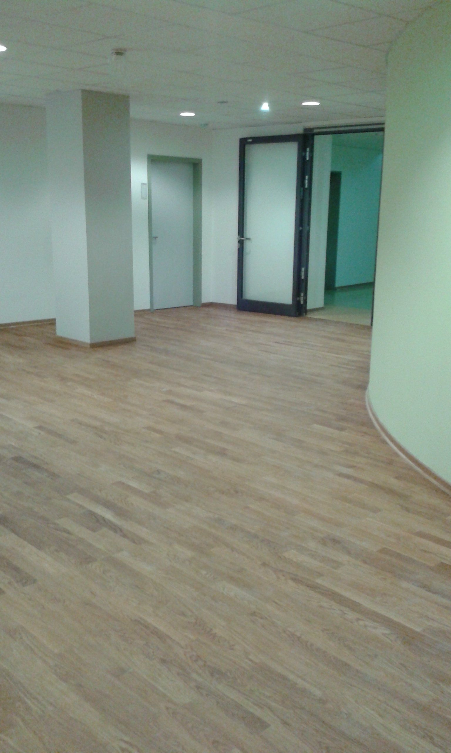 Ein leerer Büroflur mit hellem Holzboden, einer zentralen Säule, einer geschlossenen grauen Tür 