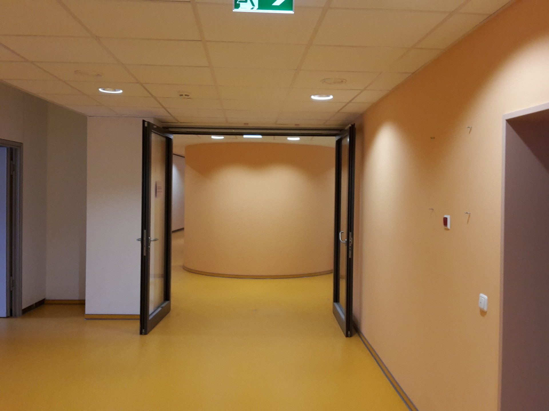 Der Blick durch offene Glastüren einen gelb gefliesten Flur entlang, der zu einer geschwungenen, pfirsichfarbenen Wand führt.