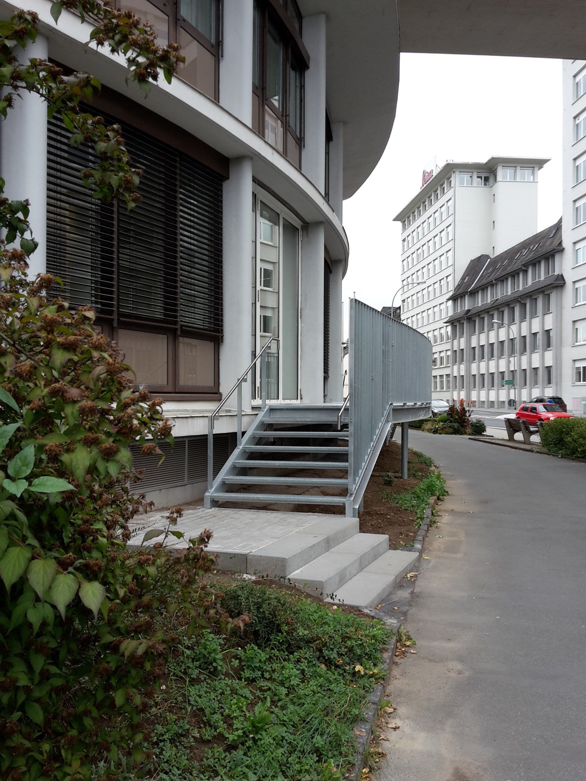 Eine Metalltreppe mit Handlauf führt zum gläsernen Eingang eines modernen Gebäudes neben einer Straße.