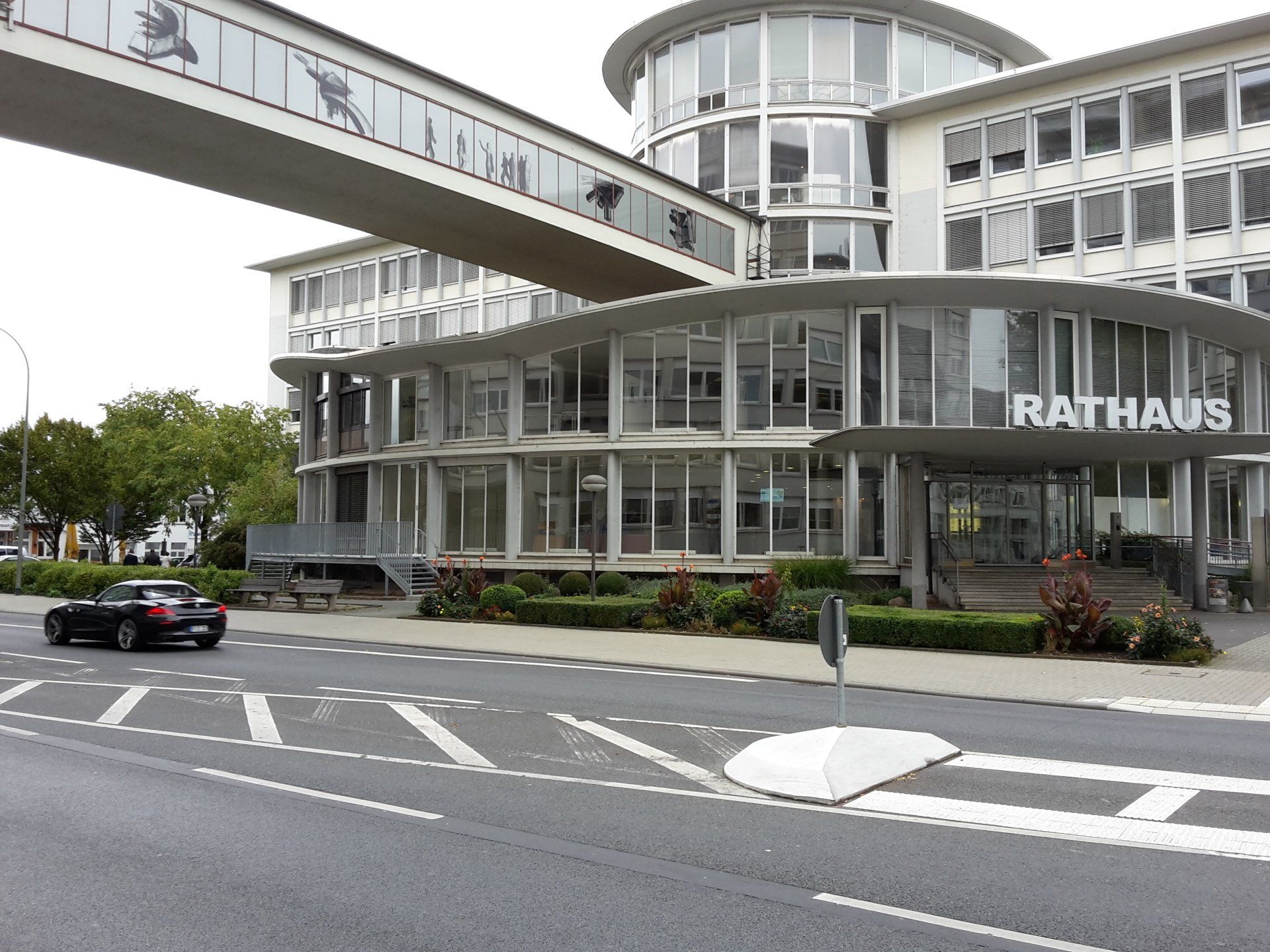 Ein modernes Rathaus mit einem großen, erhöhten Glassteg, vor dem ein schwarzes Auto auf der Straße steht.