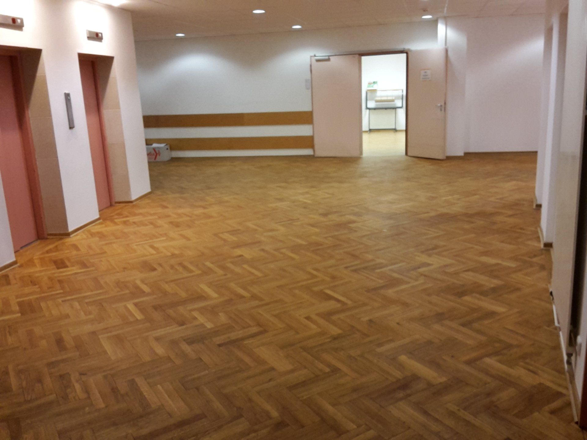 Ein Flur mit Fischgrätparkett, zwei Aufzugstüren auf der linken Seite und eine offene Tür, die in einen hellen Raum führt.