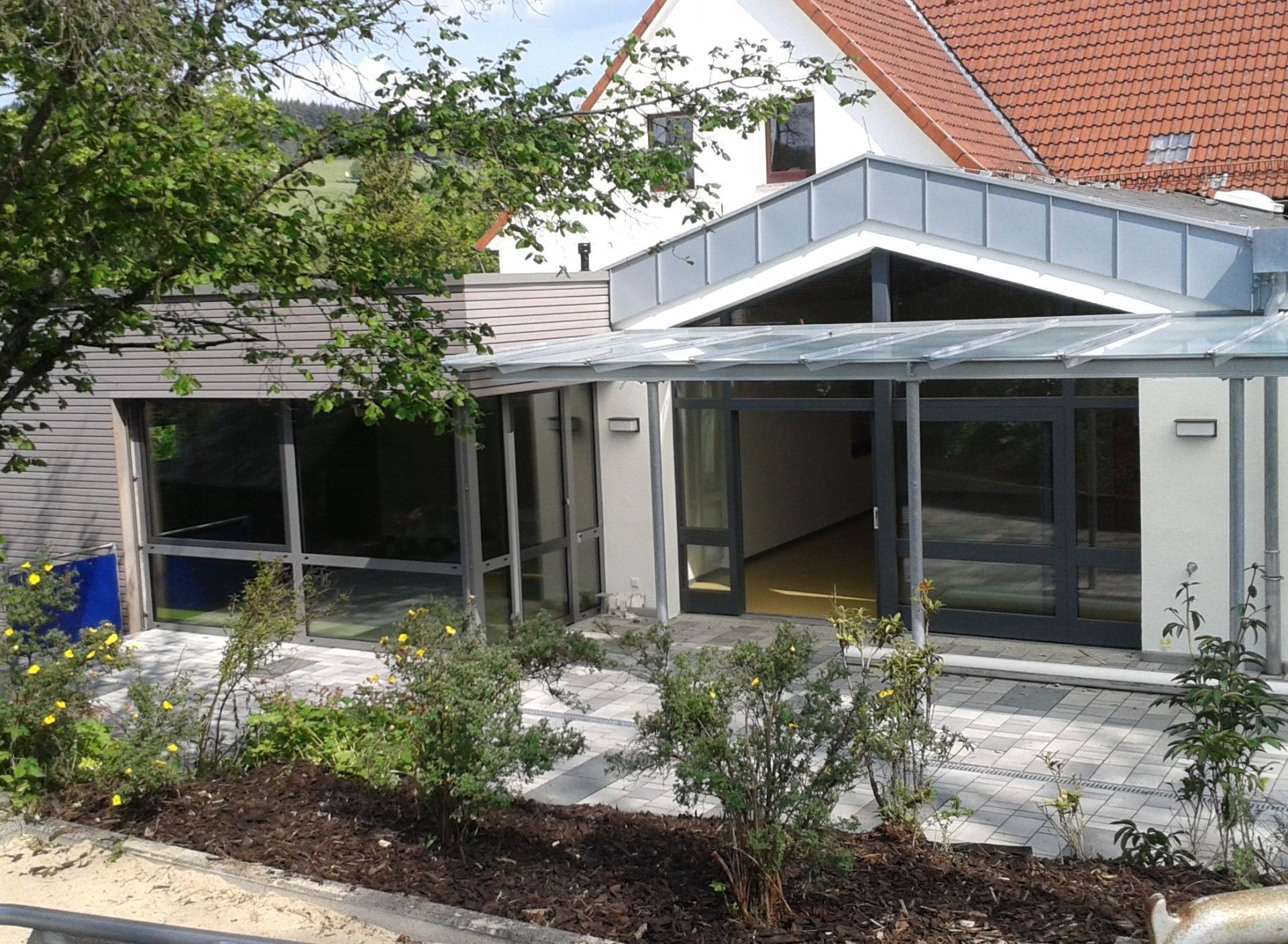 Ein moderner Gebäudeeingang mit Glasfassade, Metallmarkise und begrünter Terrasse vor einem Haus.