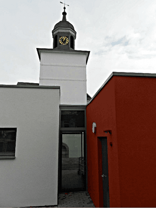 Zwischen einem modernen weißen Gebäude und einem leuchtend roten Gebäude steht ein zentraler weißer Glockenturm mit Uhr.