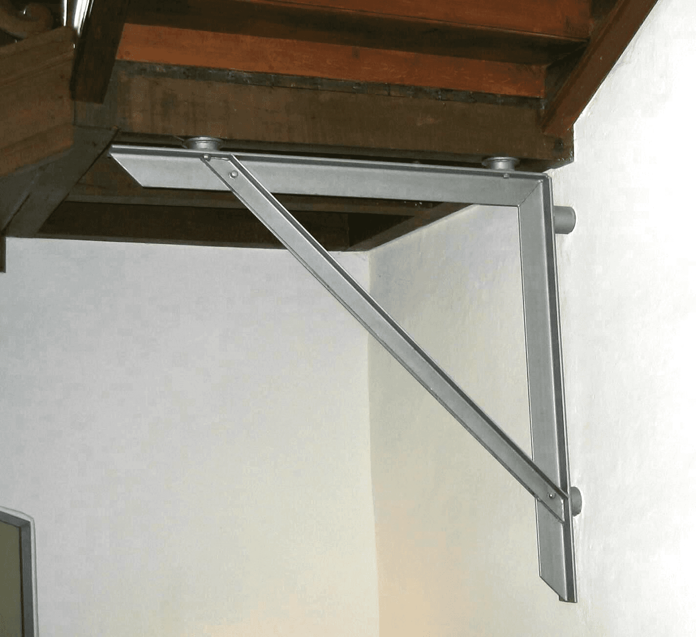Eine graue, L-förmige Metallhalterung ist an einer weißen Wand befestigt und trägt eine darüber liegende Holzkonstruktion.