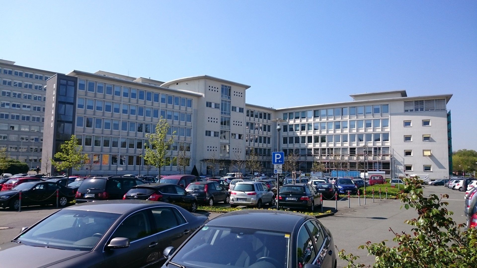 Ein großes, modernes mehrstöckiges Bürogebäude mit einem gepflasterten Parkplatz davor, auf dem zahlreiche Autos parken.