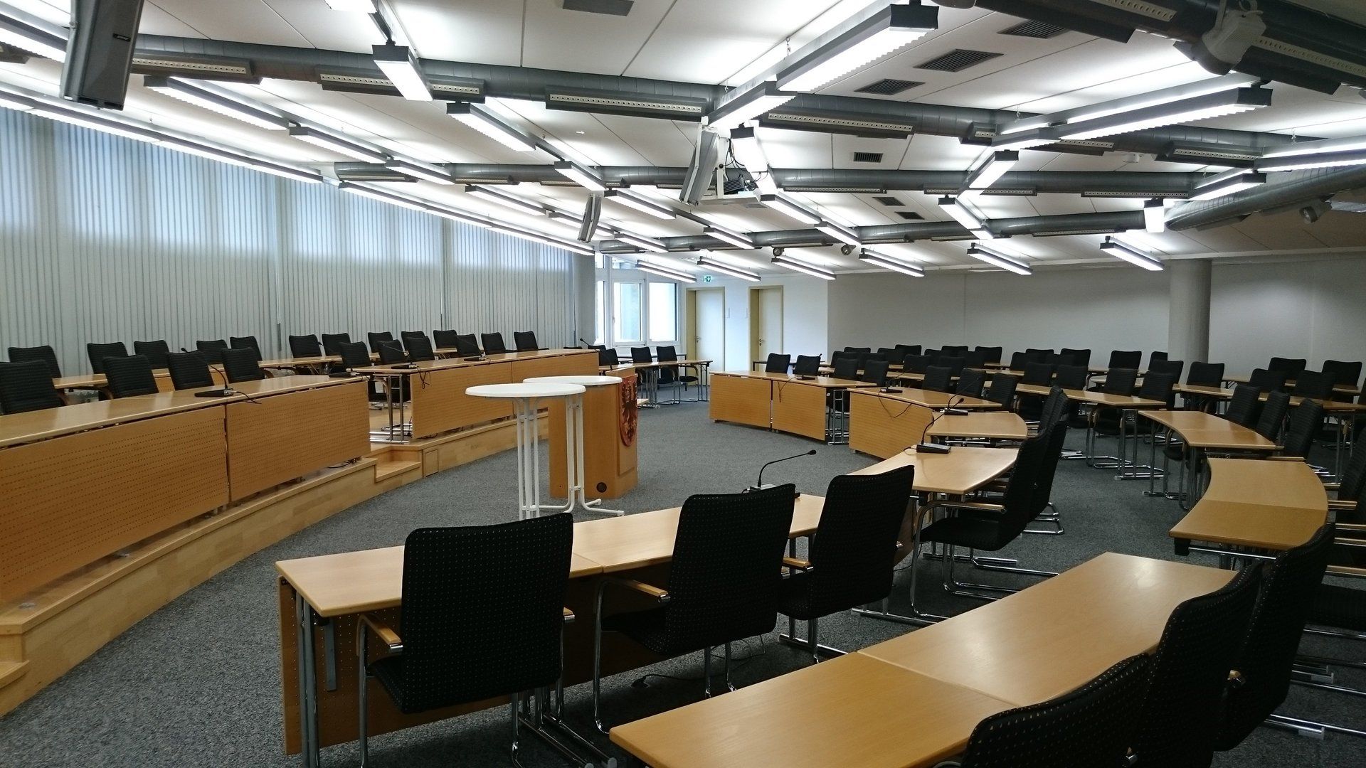 Leerer, halbrunder Besprechungsraum mit Holztischen, schwarzen Bürostühlen und moderner Deckenbeleuchtung.