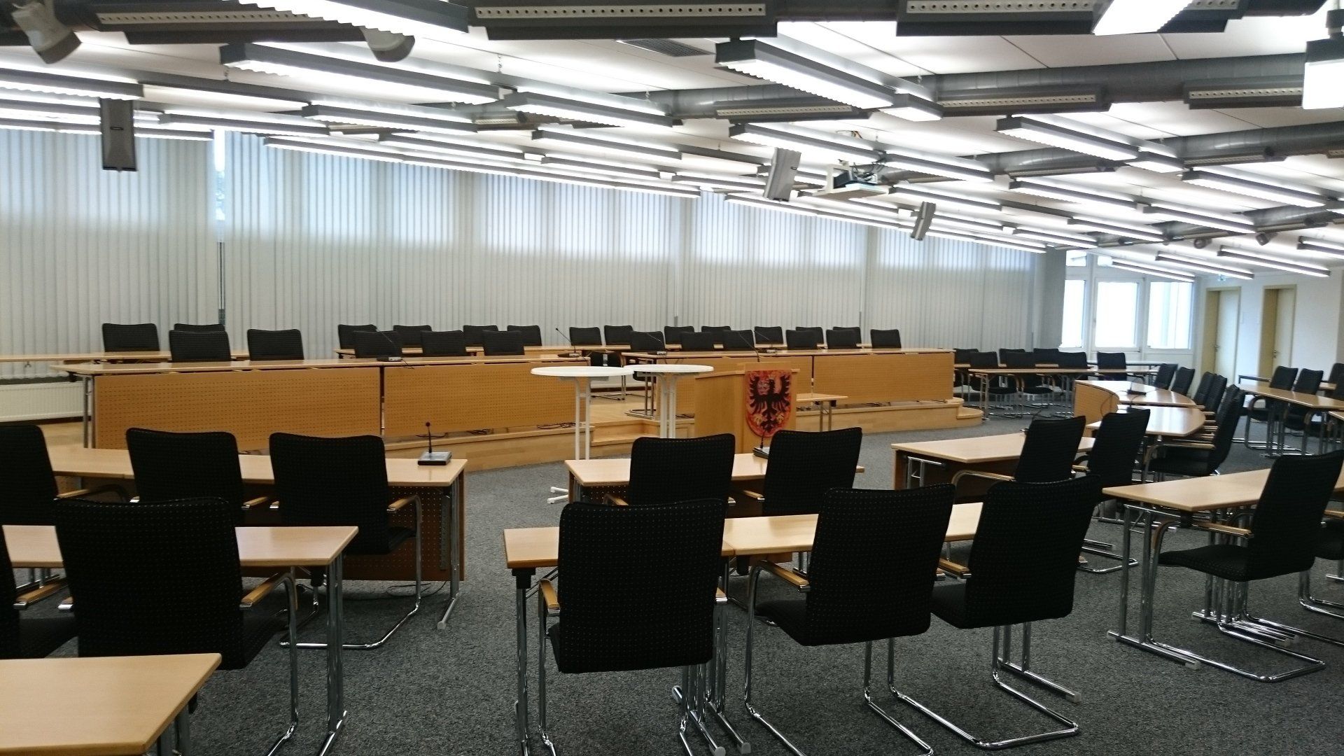 Ein formeller, leerer Versammlungssaal mit langen Holztischen, schwarzen Stühlen und einem Regierungswappen auf einem Podest.