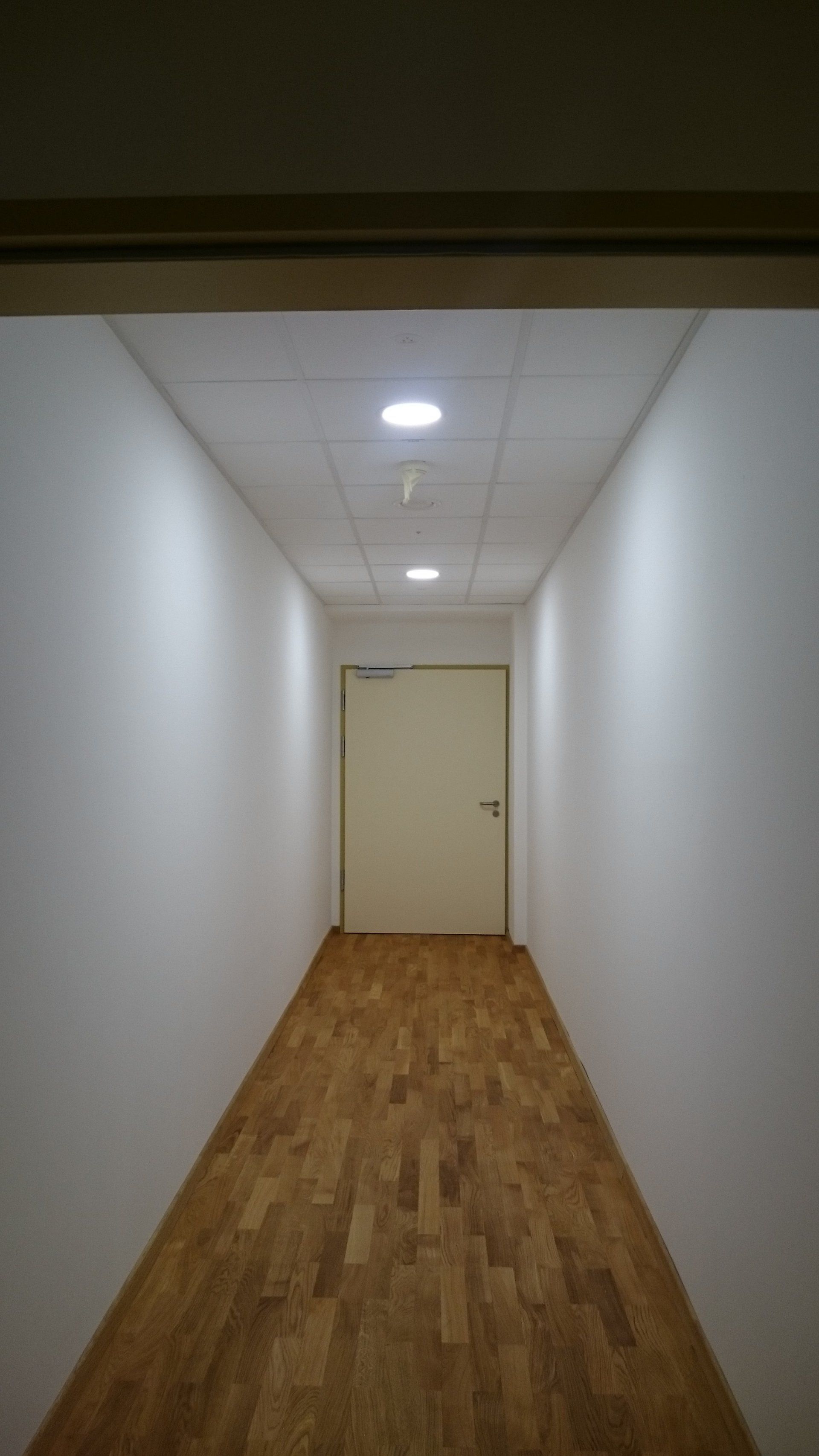 Ein langer, schmaler Flur mit weißen Wänden, Holzboden und einer geschlossenen Tür am Ende unter zwei runden Deckenleuchten.