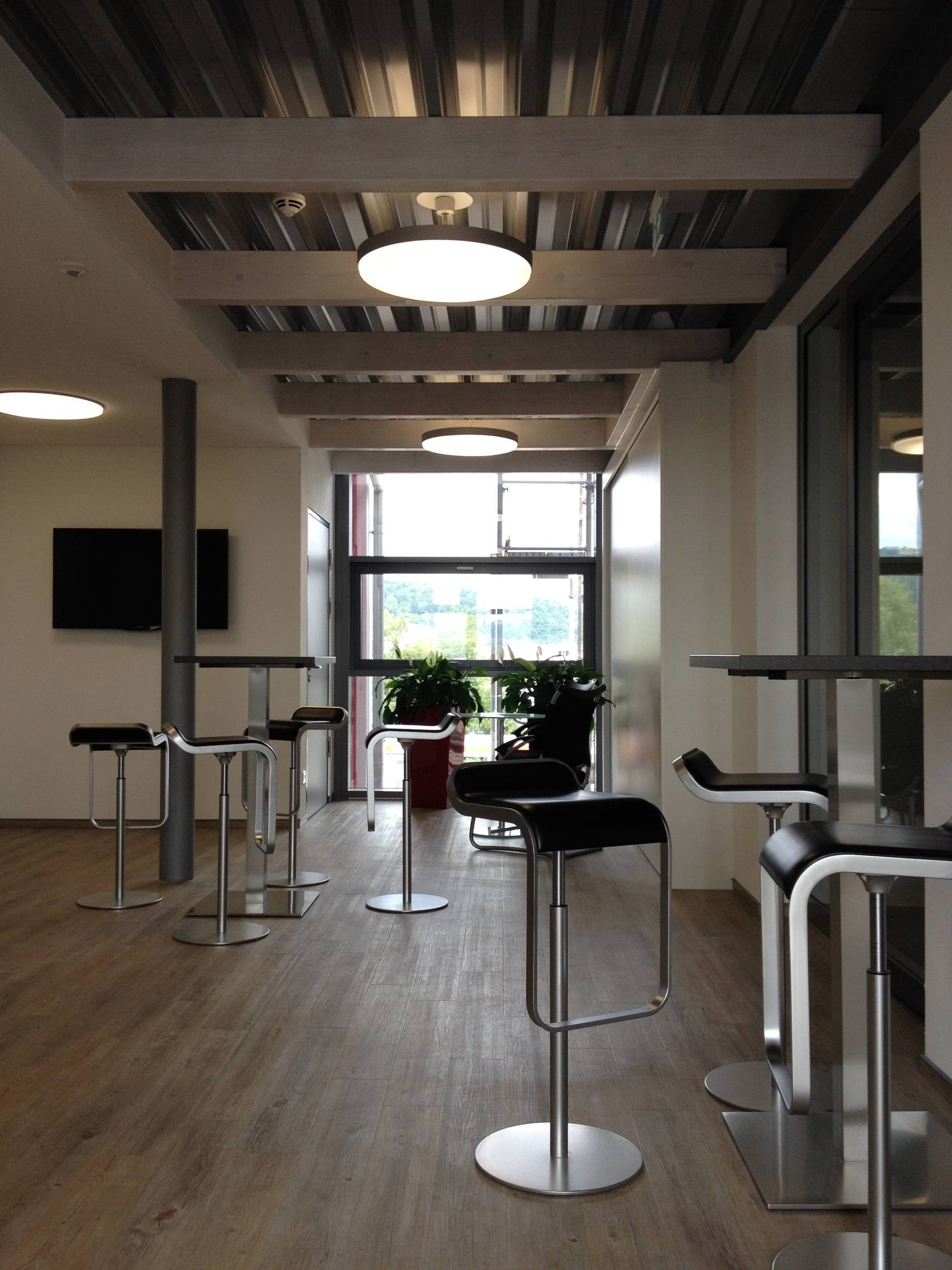 Moderner Büro-Pausenraum mit Stehtischen, Barhockern, Holzboden und Deckenleuchten mit Blick auf ein Fenster.
