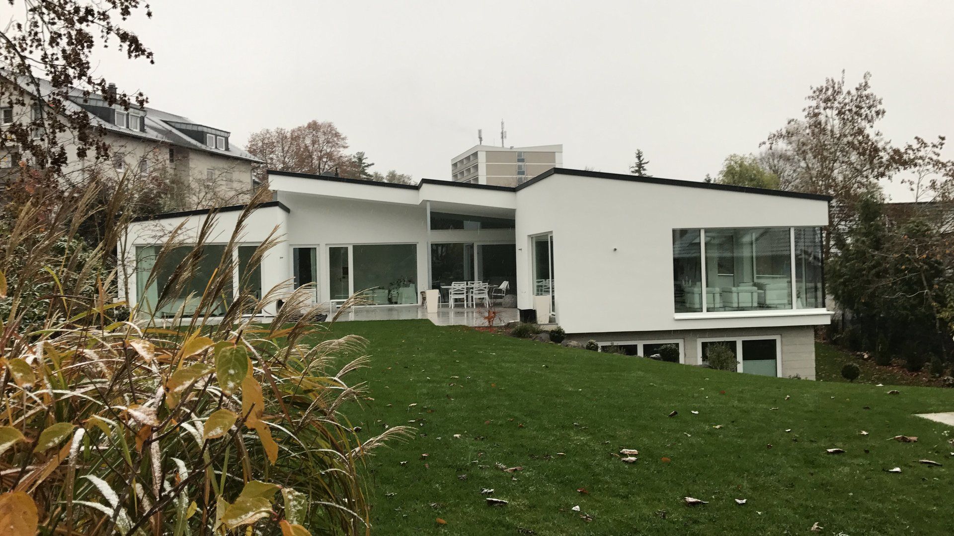 Ein modernes weißes Haus mit großen Glasfenstern überblickt einen grünen Rasen unter grauem Himmel.