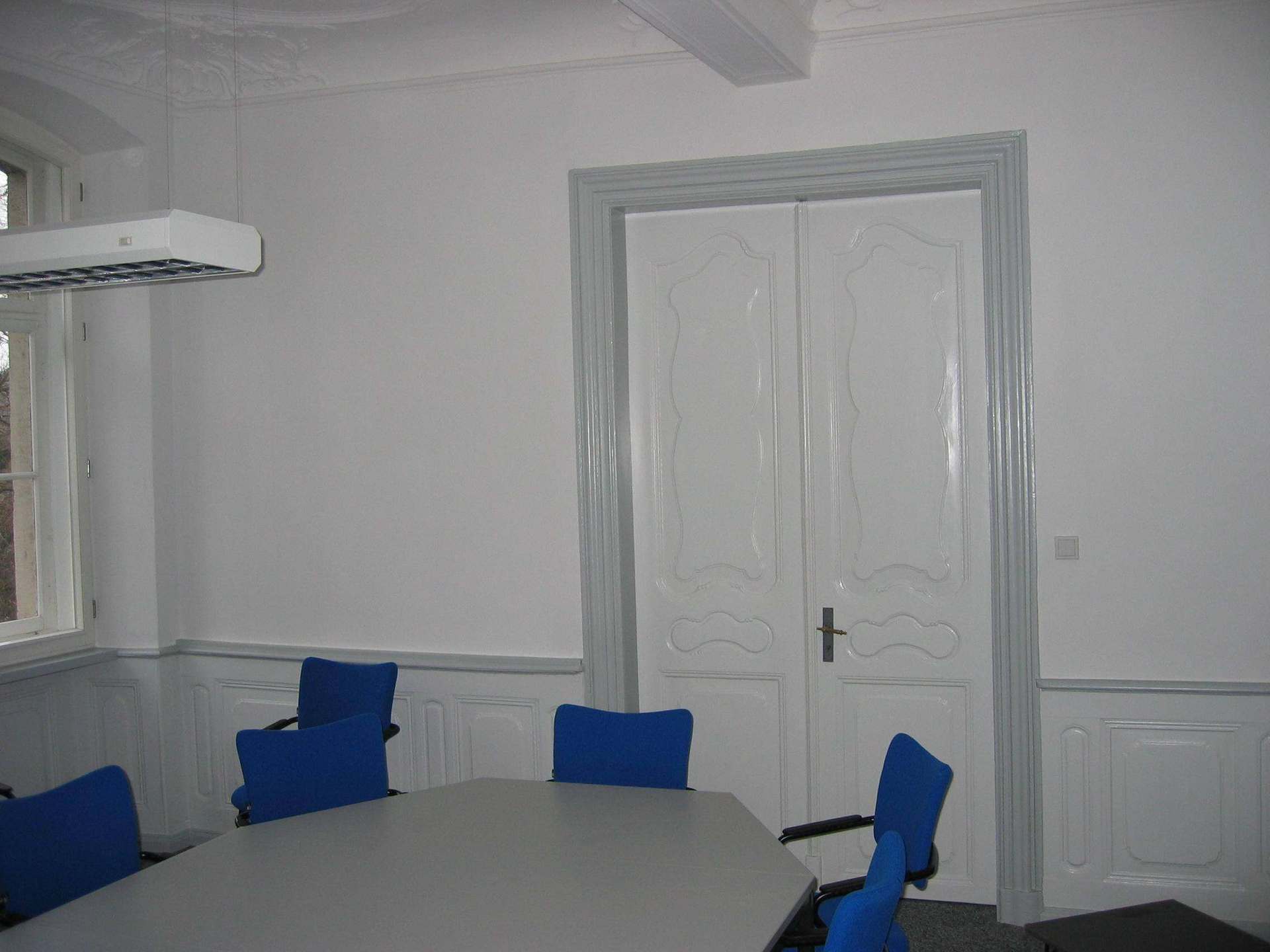 Ein Konferenztisch, umgeben von blauen Stühlen, in einem Raum mit weißen Wänden, Holzvertäfelung und großen Doppeltüren.