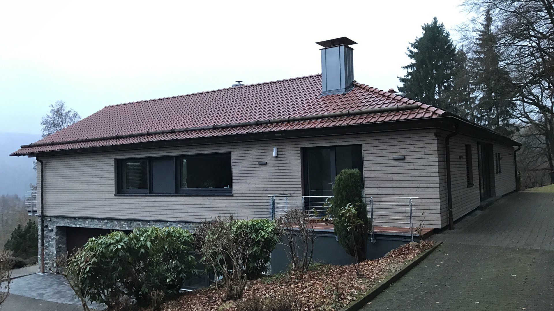 Ein eingeschossiges, helles Haus mit rotem Ziegeldach und dunklen Fensterrahmen, umgeben von Bäumen und einer 