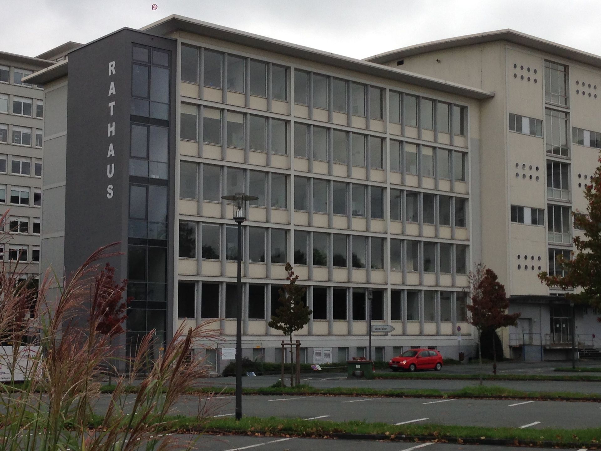 Ein mehrstöckiges graues Rathaus mit einem Raster aus Fenstern, einem davorliegenden Parkplatz und einigen Herbstbäumen.