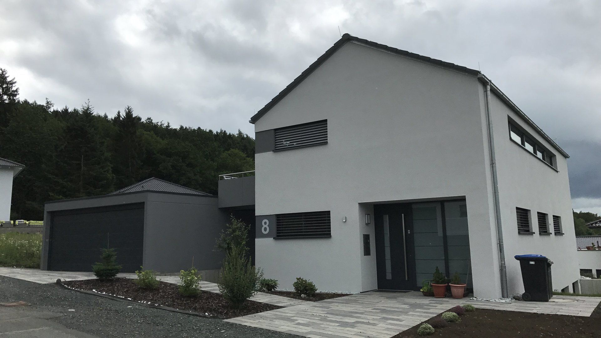 Modernes zweistöckiges Haus mit weißer Fassade, dunkelgrauer Garage und ebenem Zufahrtsweg unter bewölktem Himmel.