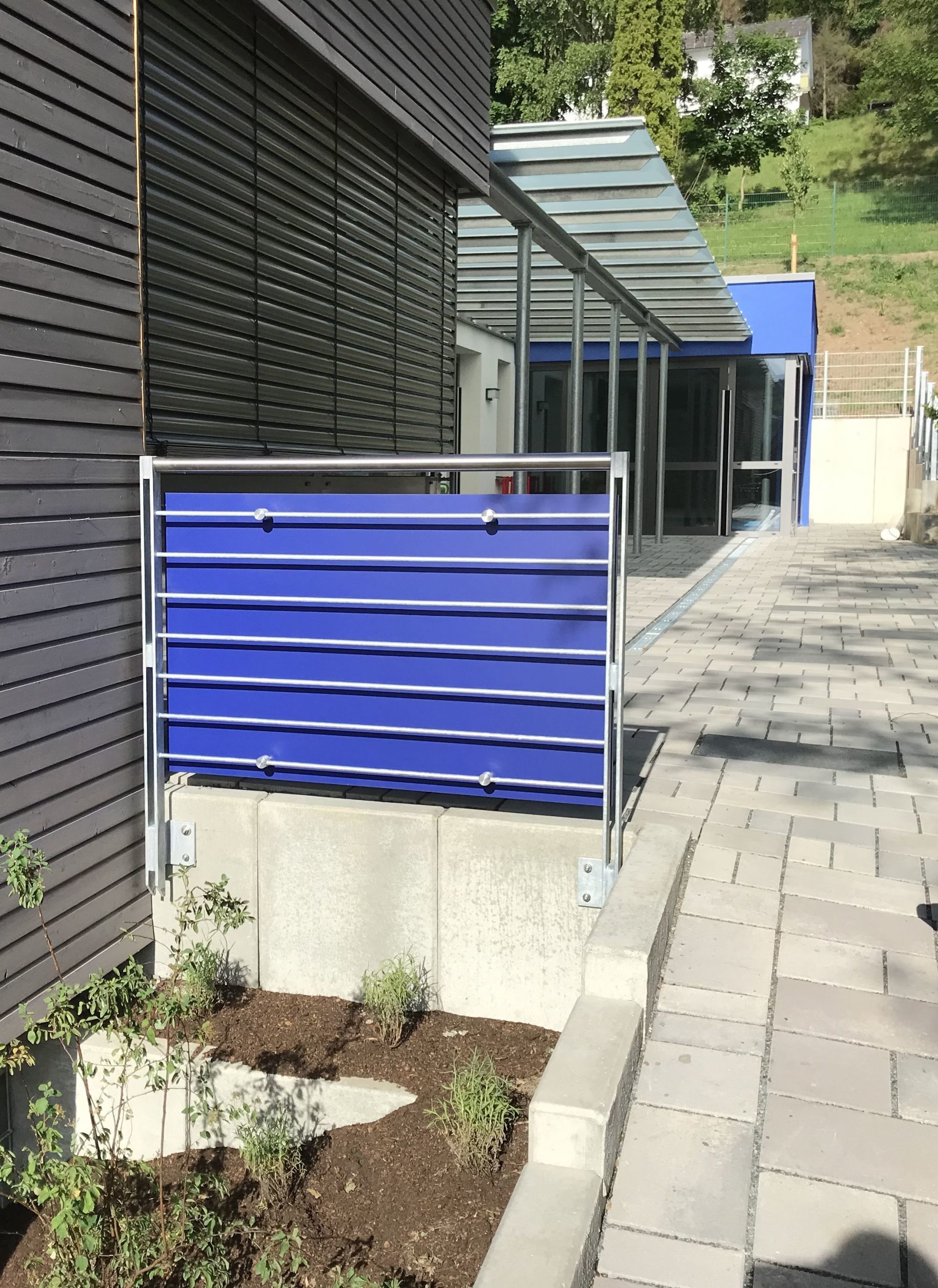 Ein blauer Sichtschutz aus Metall steht auf einem Betonfundament neben einem Holzgebäude und einem gepflasterten Weg.