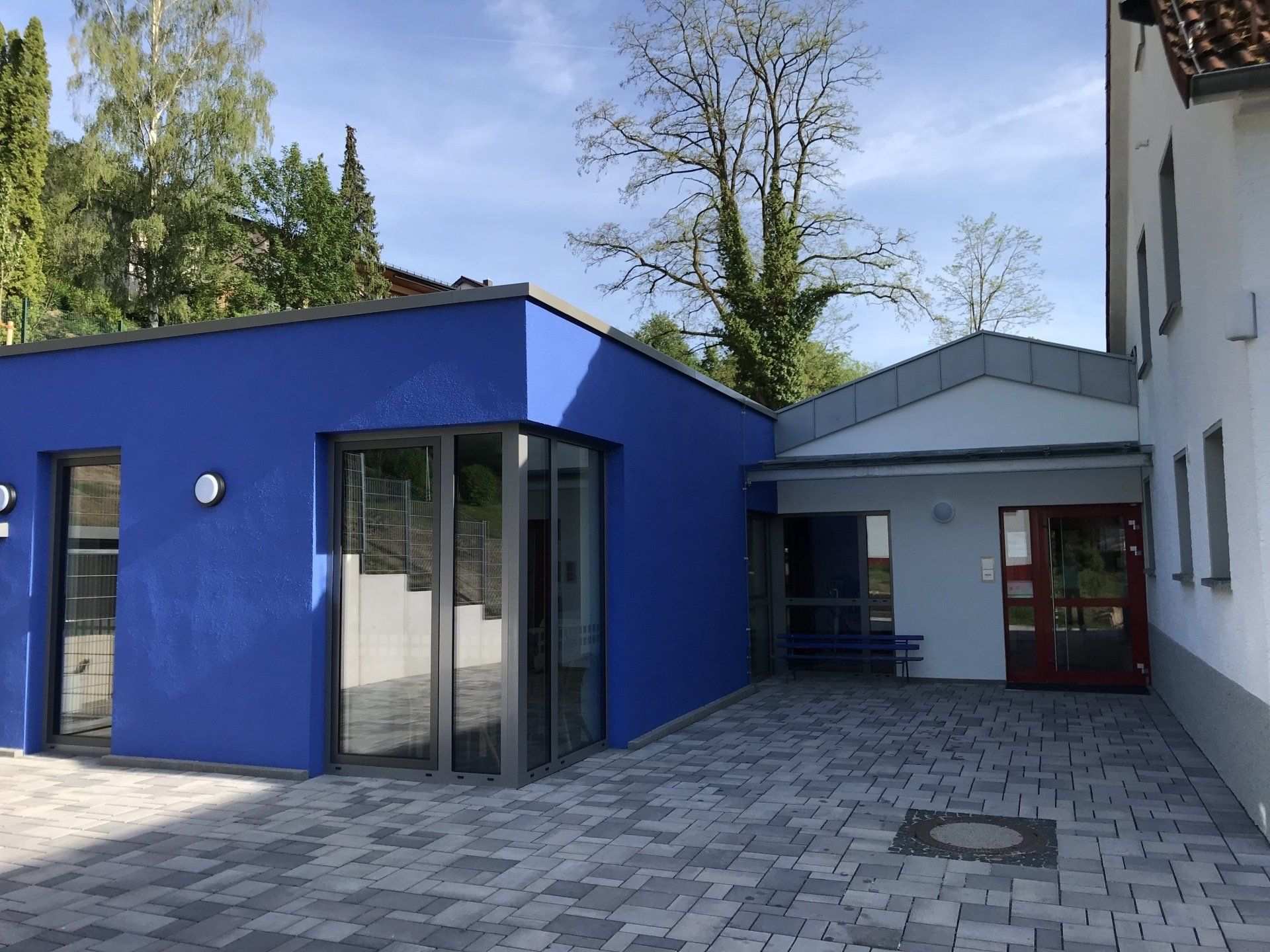 Ein moderner, hellblauer Gebäudeanbau neben einem weißen Gebäude mit gepflastertem Innenhof und roter Eingangstür.