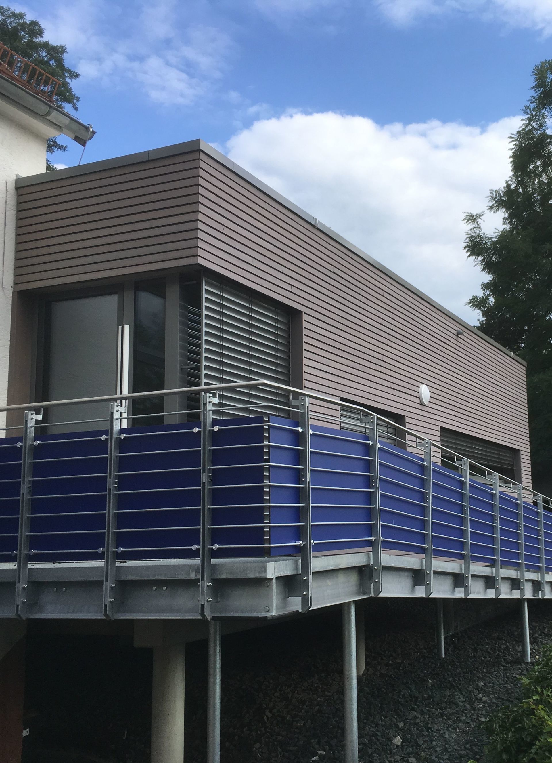 Ein modernes Gebäude mit erhöhter Terrasse, die durch eine blaue Sichtschutzwand und Metallgeländer geschützt ist,