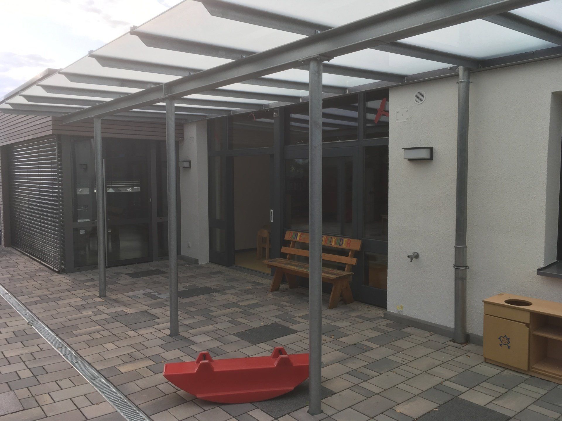 Außenterrasse eines Gebäudes mit einem metallgerahmten Glasvordach, einer Holzbank, einem roten Schaukeltier