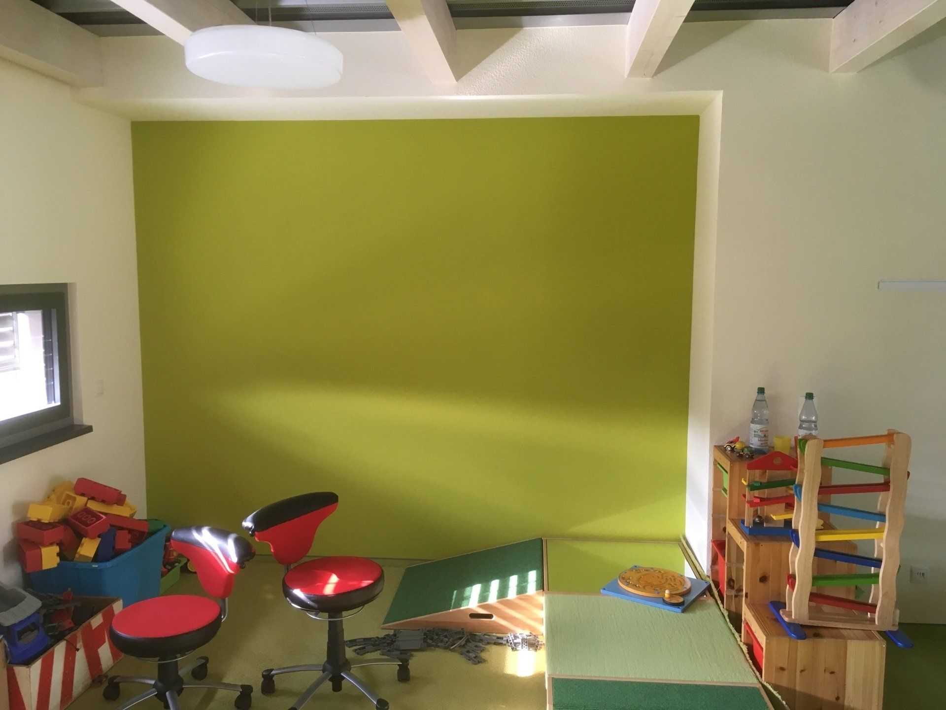 Ein hell erleuchtetes Spielzimmer mit lindgrüner Wand, roten Drehstühlen, Spielzeugkisten 