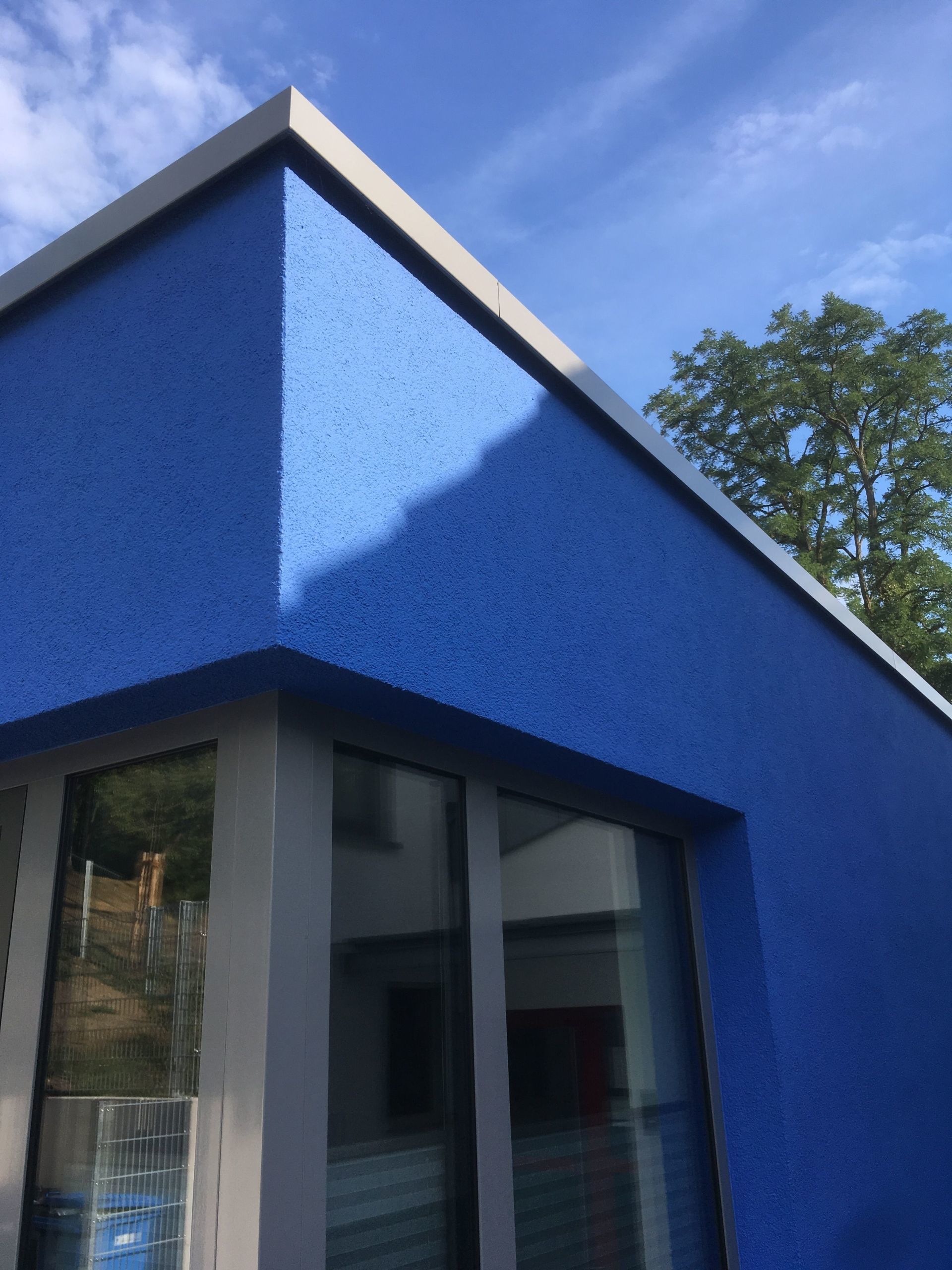 Eine Ecke eines leuchtend blauen Gebäudes mit strukturierter Fassade und grauen Zierelementen vor einem klaren blauen Himmel.