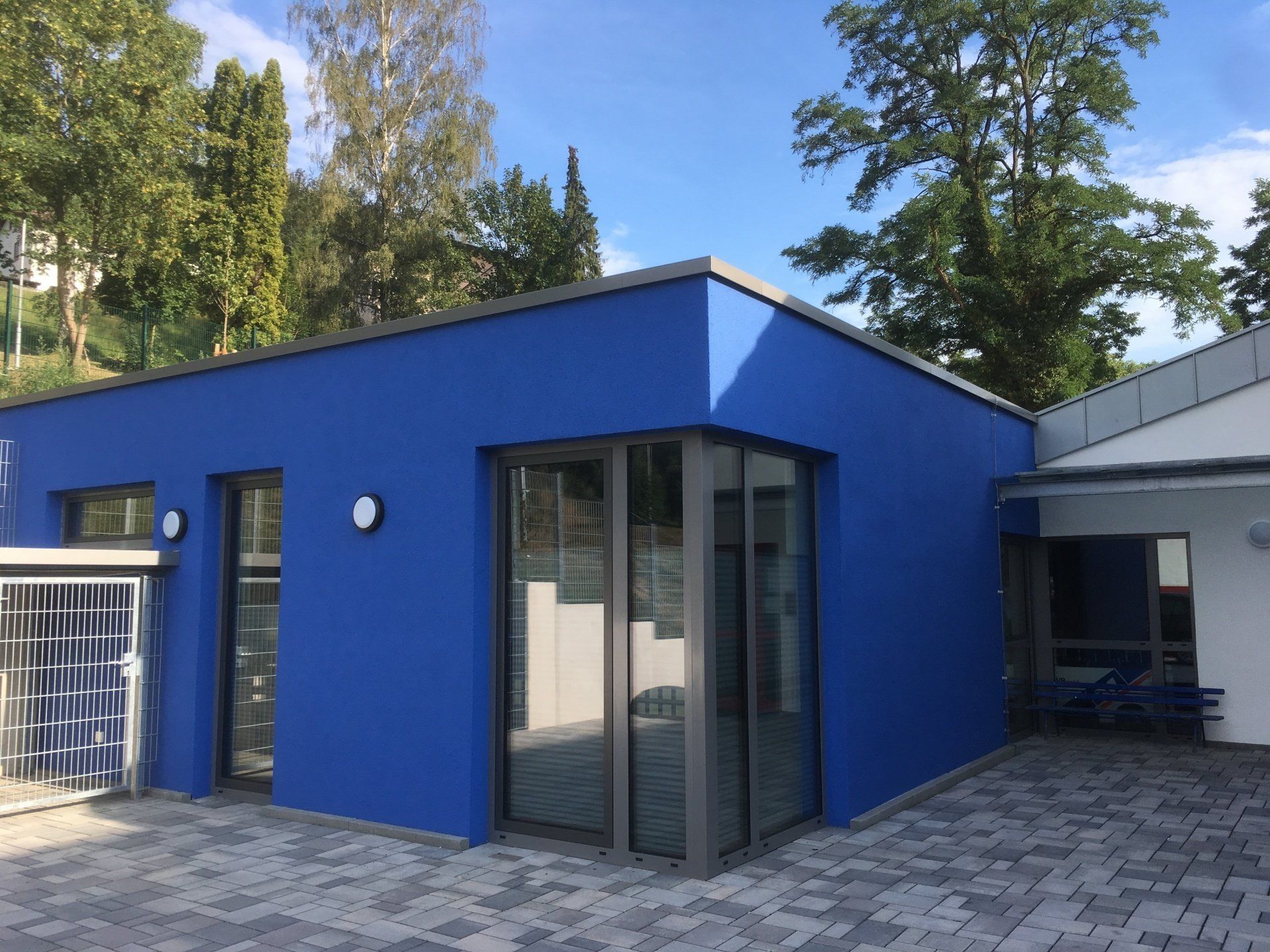 Ein modernes Gebäude mit leuchtend blauer Fassade, verglaster Ecke und gepflastertem Gehweg unter klarem, blauem Himmel.