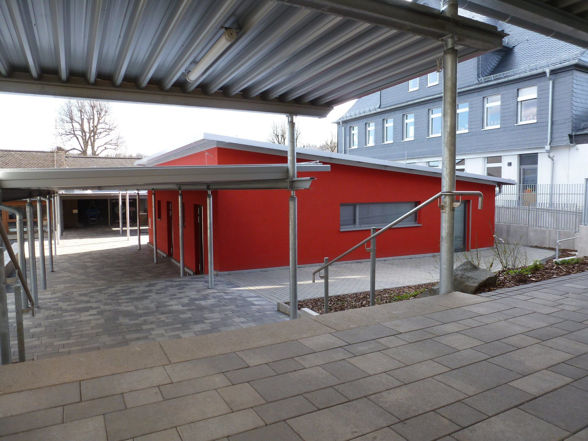Ein gepflasterter Innenhof mit einem modernen, leuchtend roten Gebäude und einem überdachten Gehweg mit Metallsäulen.