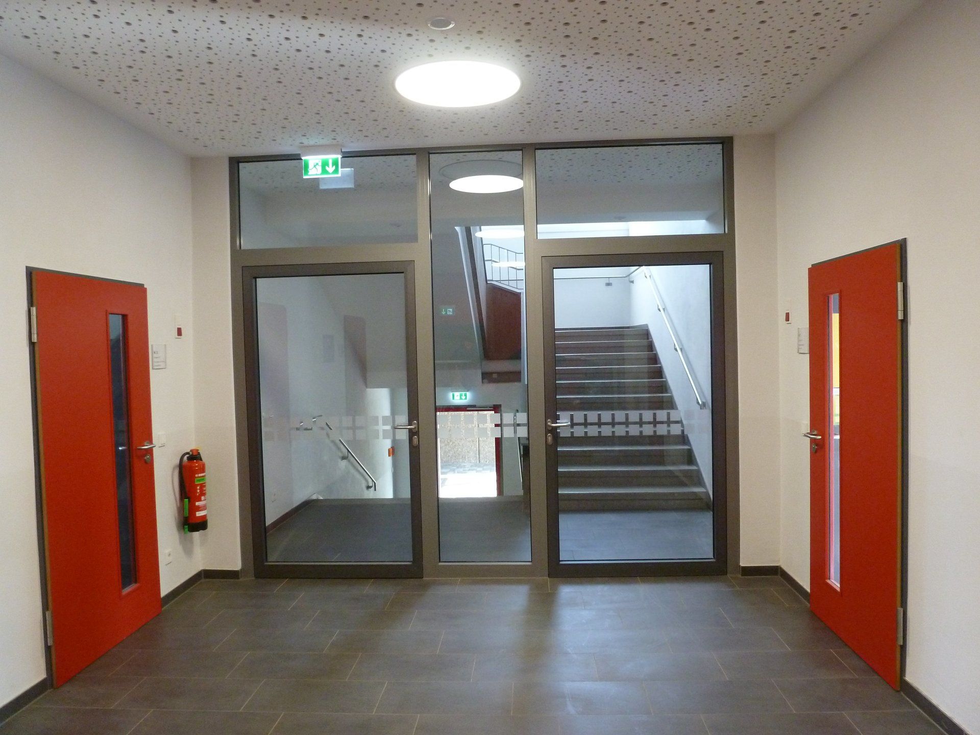 Ein Innenflur mit zwei roten Türen an den Seiten und einem zentralen, verglasten Eingang, der zu einem Treppenhaus führt.