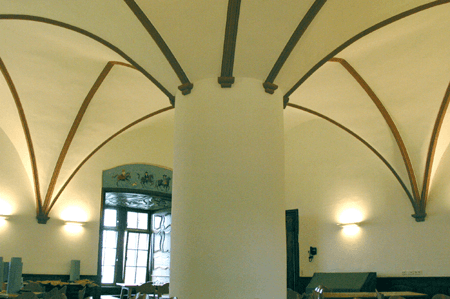 Ein heller Raum mit einer markanten Mittelsäule, weißen Gewölbedecken mit dekorativen Rippen und warmer Wandbeleuchtung.
