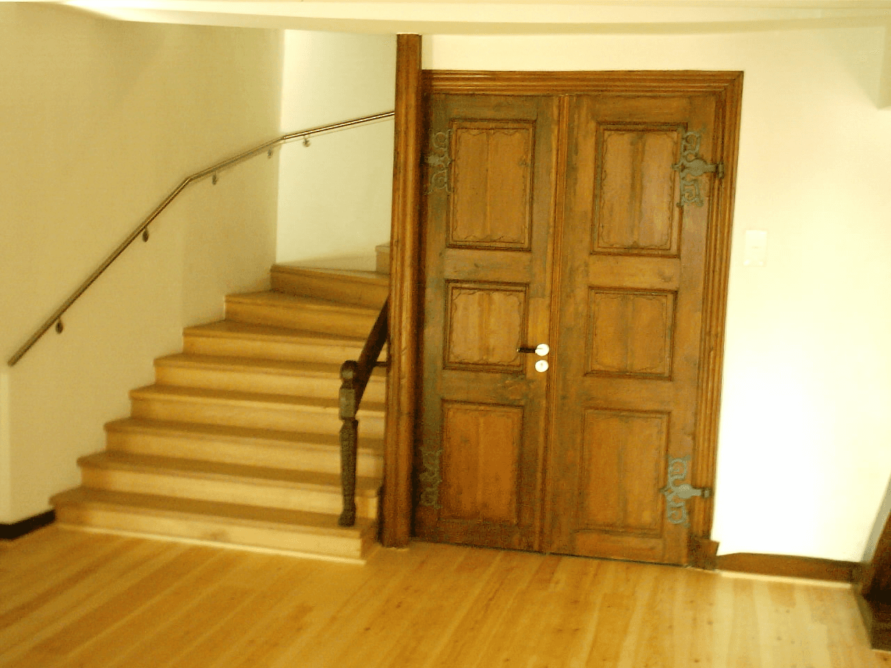 Eine Holztreppe mit Handlauf neben einer großen, doppelflügeligen Holztür in einem Raum mit hellen Wänden.