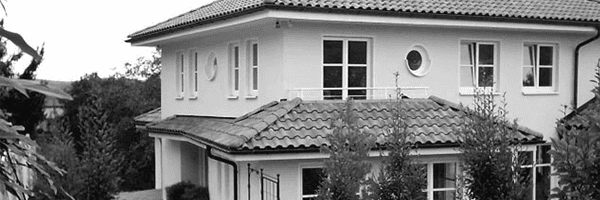 Ein zweistöckiges weißes Haus mit Ziegeldach und mehreren Fenstern, umgeben von Bäumen.