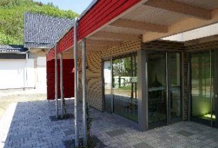 Moderner Gebäudeeingang mit roter Markise, Glastüren und hellen Holzgitterwänden über einer gepflasterten Terrasse.