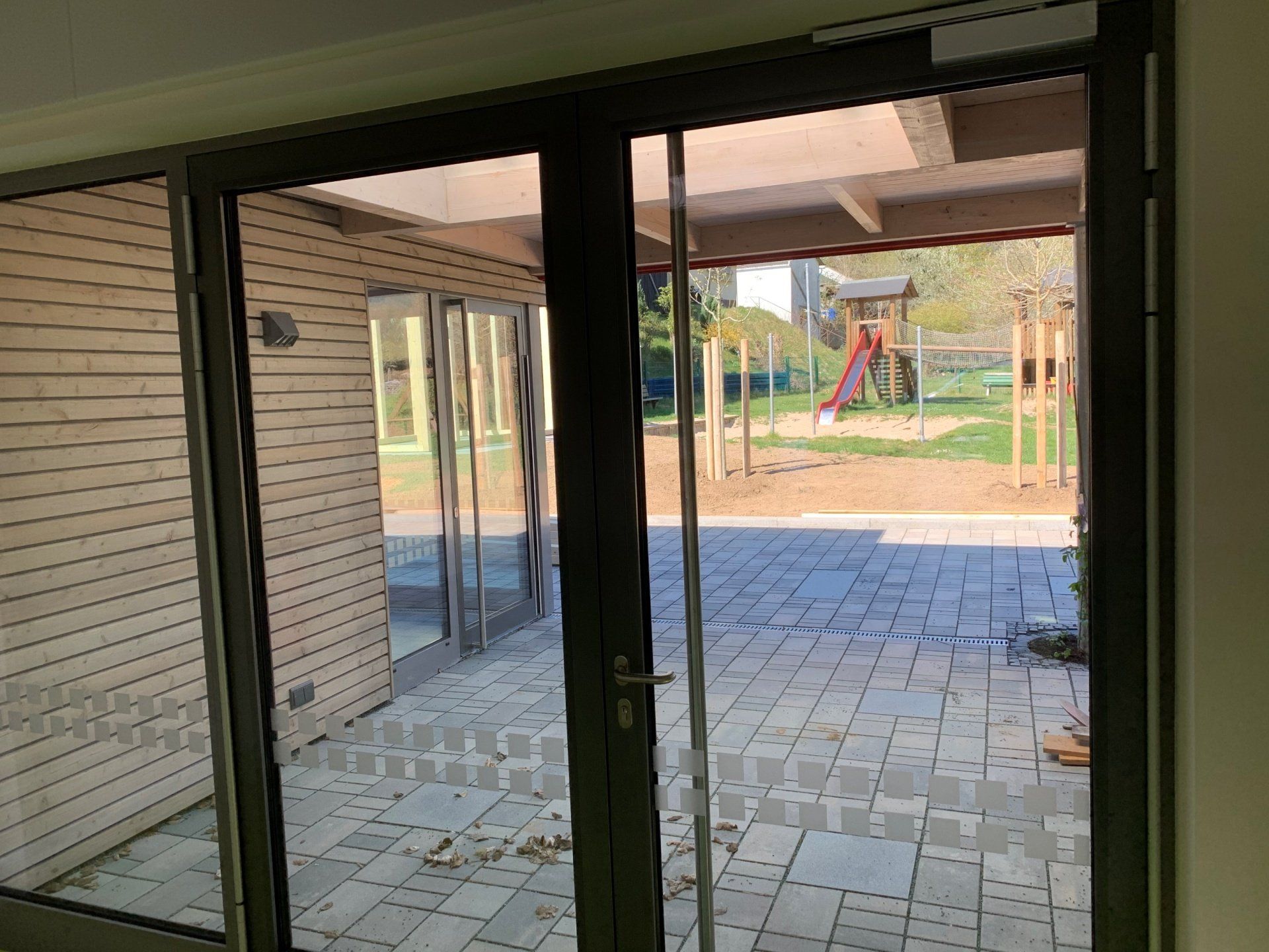 Glastüren rahmen den Blick auf eine Steinterrasse und einen Kinderspielplatz mit Rutsche in einer parkähnlichen Umgebung ein.