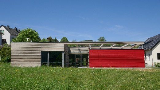 Ein modernes Gebäude mit einem Holzteil und einer leuchtend roten Wand, eingebettet in eine grasbewachsene Wiese