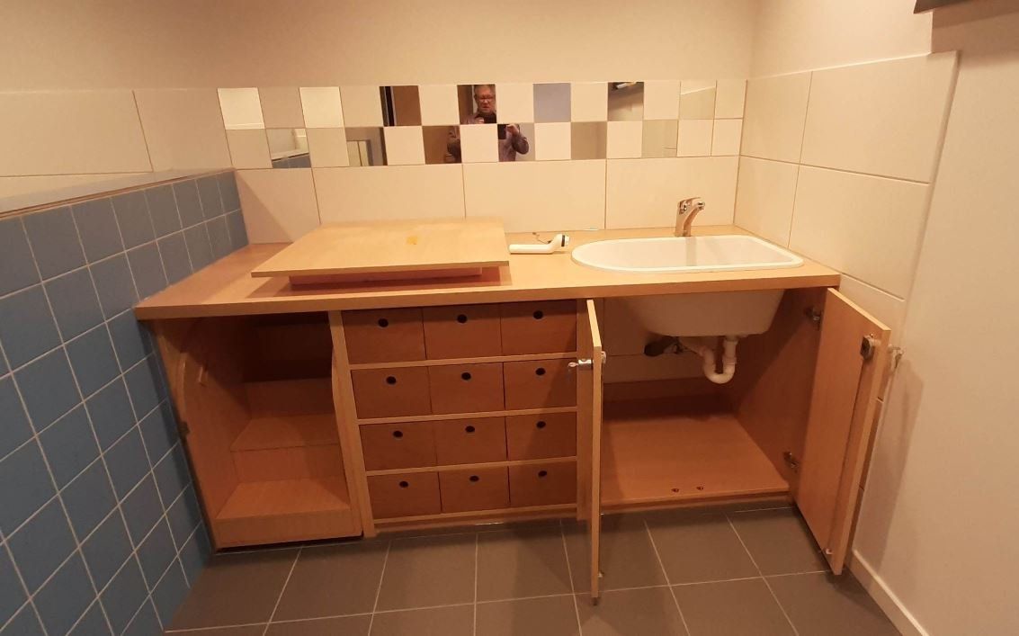 Ein hölzerner Waschtisch mit Waschbecken, offenen Schränken und einer Reihe kleiner Schubladen vor gefliesten Wänden.