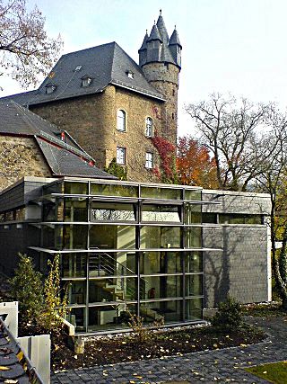 Ein moderner, verglaster Gebäudeanbau, der an einen alten steinernen Burgturm angebaut ist, umgeben von Bäumen