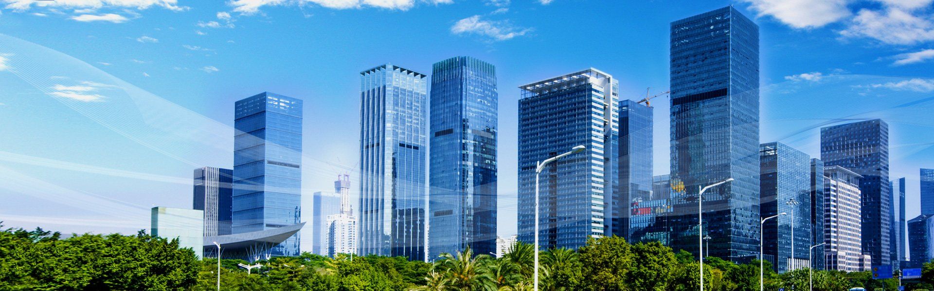 Skyscrapers against a bright blue sky with clouds, trees in the foreground.