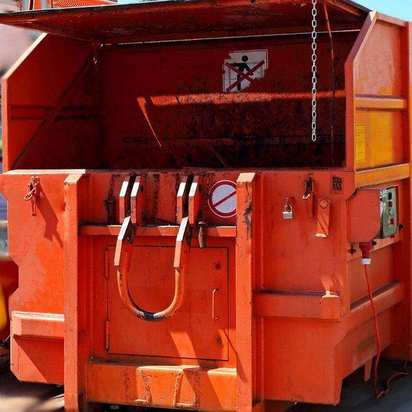 Orange industrial waste compactor with open top and warning signs.