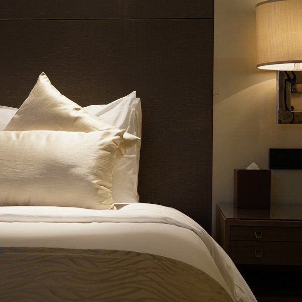 Bed with white linens and pillows, under a brown headboard, next to a lit lamp on a nightstand.