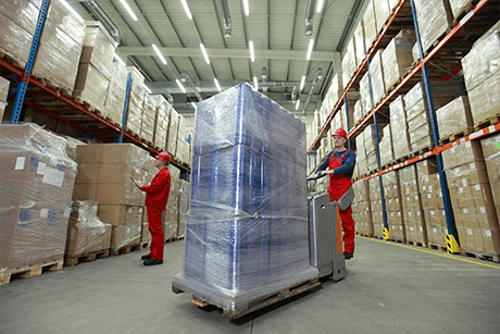 Two workers in red uniforms operate a pallet jack in a warehouse, moving a blue-wrapped pallet.